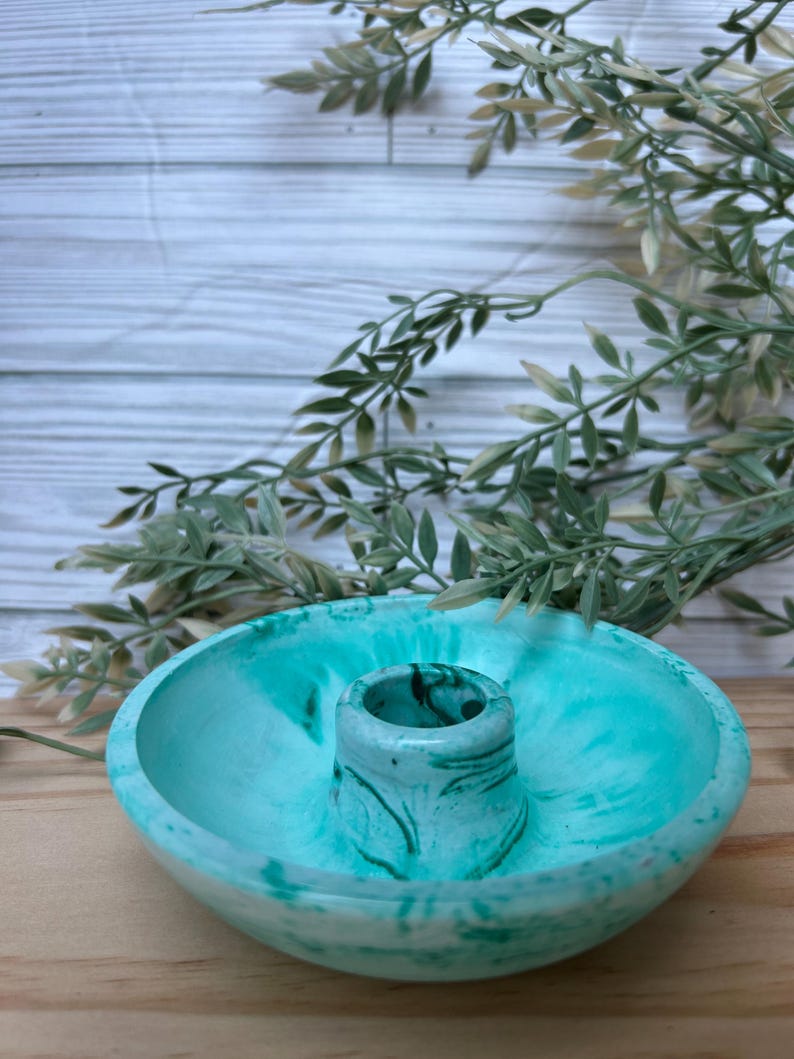 Op de afbeelding: Een teal en wit gemarmerde betonnen kandelaar. De kandelaar heeft een cirkelvormige basis met een verhoogd midden voor de kaars. De achtergrond omvat groen en een wit houten oppervlak.