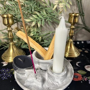 May include: A circular gray and white marbled concrete tray featuring a central holder for incense sticks, a small circular indentation for a black tea light candle, and a cylindrical socket containing a single white taper candle.