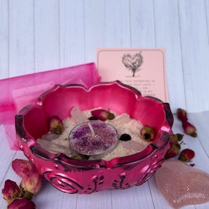 Op de afbeelding: Roze keramische kom met een bloemmotief, gevuld met wit zand, rozenkwartskristallen en een kleine roze en paarse glitterkaars. De kom is omringd door gedroogde rozenknoppen en een roze hartvormig kristal.