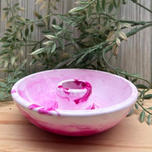 Op de afbeelding: Een roze en witte gemarmerde keramische kom met een centrale cilindrische houder. De kom heeft een afgeronde vorm en is versierd met wervelingen van roze en wit. De achtergrond toont groen en een houten oppervlak.