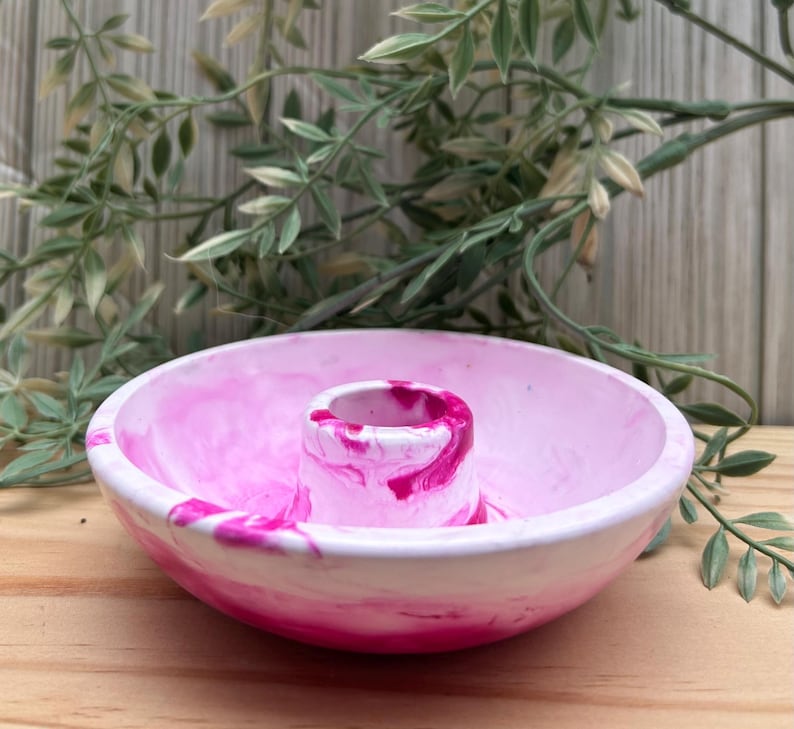 Op de afbeelding: Een roze en witte gemarmerde keramische kom met een centrale cilindrische houder. De kom heeft een afgeronde vorm en is versierd met wervelingen van roze en wit. De achtergrond toont groen en een houten oppervlak.