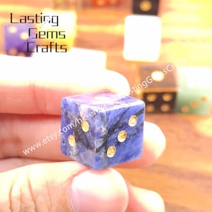 May include: A close-up of a blue and gold dice. The dice is made of a dark blue stone with gold-colored dots. The dice is being held by a person. The text "Lasting Gems Crafts" and a website address are visible.