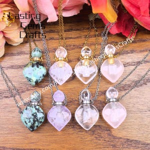 May include: A collection of gemstone and glass perfume bottle necklaces. The bottles are various shapes and colors, including green, purple, clear, and pink. Each bottle hangs from a silver or gold chain. The background is a wooden surface.