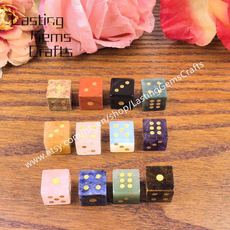 May include: A collection of gemstone dice in various colors, including pink, blue, green, and black, with gold-colored dots. The dice are arranged on a wooden surface, with the text "Lasting Gems Crafts" and a website address visible.