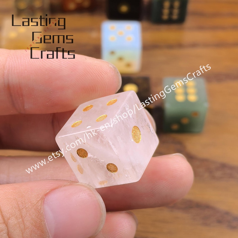 May include: A clear, quartz dice with gold-colored dots is held in a hand. The dice is in focus, with other dice in the background. The text "Lasting Gems Crafts" and a website address are visible.