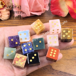 May include: A collection of gemstone dice in various colors, including purple, blue, green, and orange. Each die features gold-colored dots. The dice are held in a hand, with a blurred background of flowers and text.