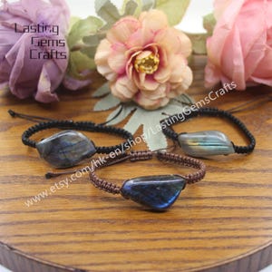 Könnte beinhalten: Drei Armbänder mit schwarzem Band und einem dunkelblauen Labradorit-Stein. Die Armbänder liegen auf einer Holzoberfläche mit rosa Blumen im Hintergrund. Der Text "www.etsy.com/hk-en/shop/LastingGemsCrafts" ist im Bild sichtbar.