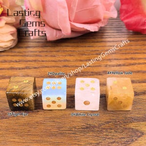 May include: Four gemstone dice: Tiger Eye, Opalite, White Crystal, and Yellow Jade. Each die features gold-colored dots. The dice are displayed on a wooden surface, with the shop name "Lasting Gems Crafts" visible.