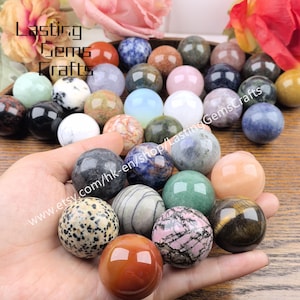 Pode incluir: Uma coleção de esferas de pedras preciosas polidas em várias cores e padrões. As esferas são seguradas em uma mão, com outras dispostas ao fundo. A imagem inclui o texto "Lasting Gems Crafts" e um endereço de site.