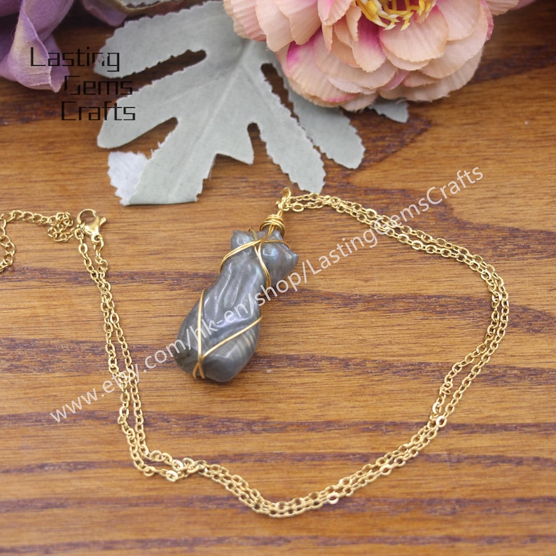 May include: A gold chain necklace with a grey labradorite stone pendant wrapped in gold wire. The pendant is shaped like a human torso. The necklace is on a wooden surface.