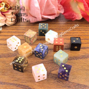 May include: A collection of gemstone dice in various colors, including pink, blue, green, and black, with gold-colored dot markings. The dice are arranged on a wooden surface, with the text "Lasting Gems Crafts" visible in the background.
