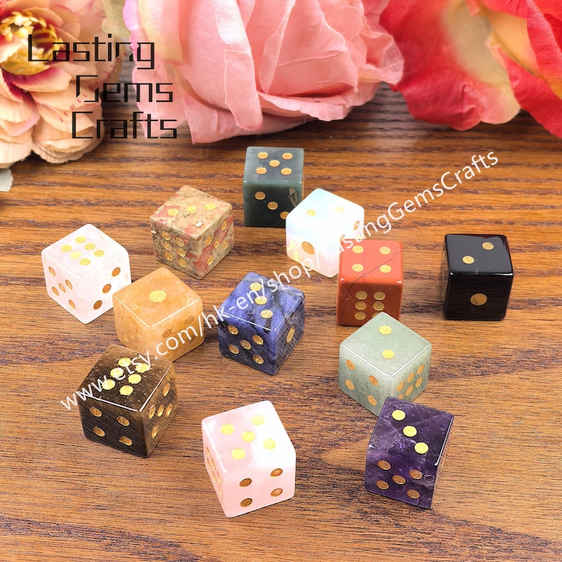 May include: A collection of gemstone dice in various colors, including pink, blue, green, and black, with gold-colored dot markings. The dice are arranged on a wooden surface, with the text "Lasting Gems Crafts" visible in the background.
