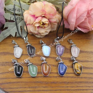 May include: A set of ten gemstone pendants with snake head accents. The pendants are made of various gemstones, including amethyst, rose quartz, tiger's eye, and lapis lazuli. Each pendant is attached to a black cord.