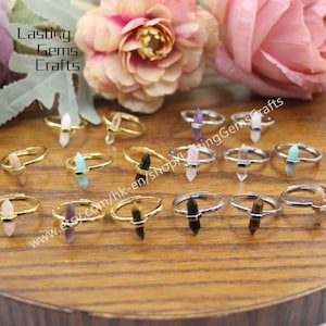May include: A variety of gemstone rings with different colored stones, including clear, pink, green, purple, black, and brown. The rings are made of gold and silver and are displayed on a wooden surface. www.etsy.com/hk-en/shop/LastingGemsCrafts
