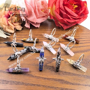 Puede incluir: Una colección de anillos de piedras preciosas con engastes plateados. Los anillos presentan varias formas y colores de cristal, incluyendo transparente, rosa, morado y negro. El fondo incluye flores y el texto "Lasting Gems Crafts".