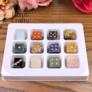 May include: A set of twelve gemstone dice in a white tray. Each die is a different color and pattern, with gold-colored dots. The dice are arranged in a grid, showcasing the variety of materials and designs. The text "Lasting Gems Crafts" is visible.