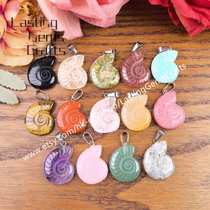 May include: A collection of gemstone pendants in various colors, including black, white, green, red, and pink. Each pendant is shaped like a spiral shell and has a silver-colored bail. The pendants are displayed on a wooden surface.