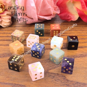 May include: A collection of gemstone dice in various colors, including pink, blue, green, and brown, with gold-colored dot markings. The dice are arranged on a wooden surface, with the text "Lasting Gems Crafts" and a website address visible.