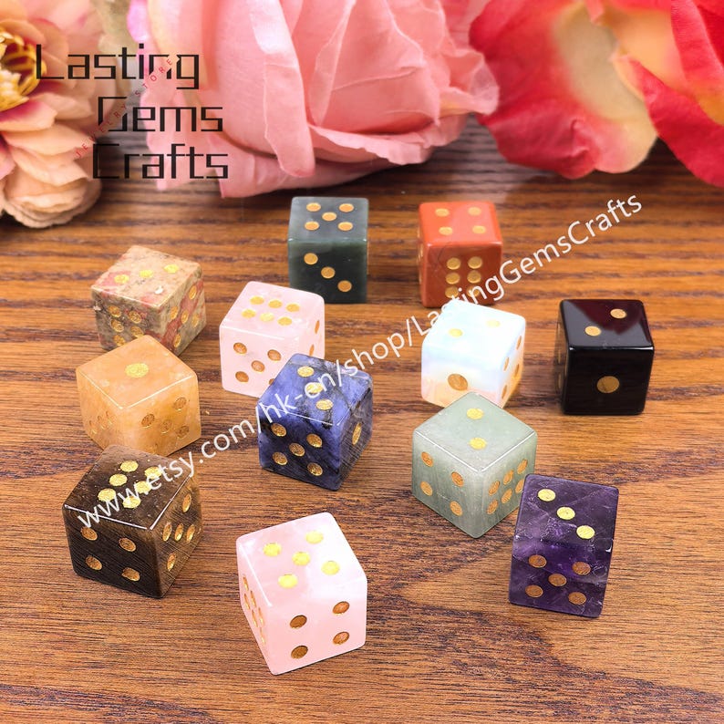 May include: A collection of gemstone dice in various colors, including pink, blue, green, and brown, with gold-colored dot markings. The dice are arranged on a wooden surface, with the text "Lasting Gems Crafts" and a website address visible.