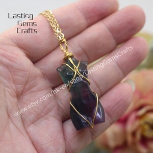 May include: A gold chain necklace with a fluorite pendant shaped like a human torso. The pendant is wrapped in gold wire and has a purple, green, and black color.