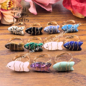 May include: A collection of gemstone fish rings with gold-colored wire detailing. The rings feature various colors and patterns, including black, blue, green, and white. The rings are displayed on a wooden surface. The text "Lasting Gems Crafts" is visible.