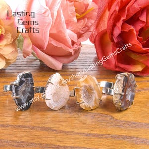 May include: Four gemstone rings with silver bands are displayed. The rings feature unique, irregularly shaped stones in shades of black, white, and brown. The background includes pink and red flowers, and the text "Lasting Gems Crafts".