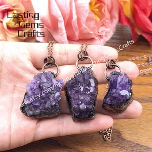 May include: Three amethyst crystal pendants with dark metal chains. The raw, purple crystals are mounted on a dark base. The image includes the text "Lasting Gems Crafts" and a website address.