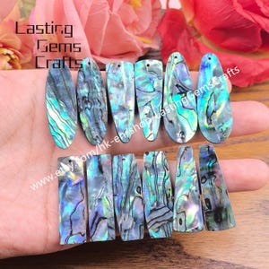 May include: A collection of iridescent abalone shell pendants in various shapes, including oval, teardrop, and rectangular. The pendants display a mix of blue, green, and purple hues with natural patterns. The text "Lasting Gems Crafts" is visible.