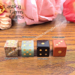 May include: Four gemstone dice: Red Unakite, Aventurine, Obsidian, and Red Jasper. Each die has gold-colored dots. The dice are displayed on a wooden surface with the text "Lasting Gems Crafts".