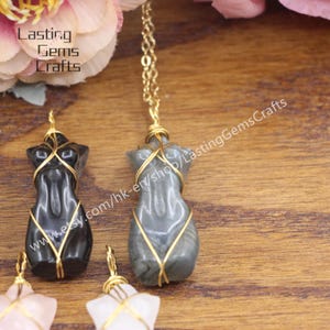 May include: Two wire-wrapped pendants, one black and one gray, shaped like a human torso. The pendants are on a wooden surface with a gold chain necklace in the background. The text "www.etsy.com/hk-en/shop/LastingGemsCrafts" is visible in the image.