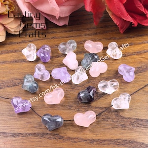May include: A collection of heart-shaped gemstones in various colors, including clear, pink, purple, and gray. Some are strung on a black cord. The Etsy shop name "Lasting Gems Crafts" is visible in the image.