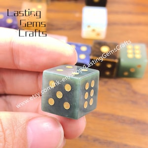May include: A green gemstone die with gold-colored dots is held in a hand. The die is in focus, with other dice of various colors blurred in the background. The text "Lasting Gems Crafts" and a website address are visible.