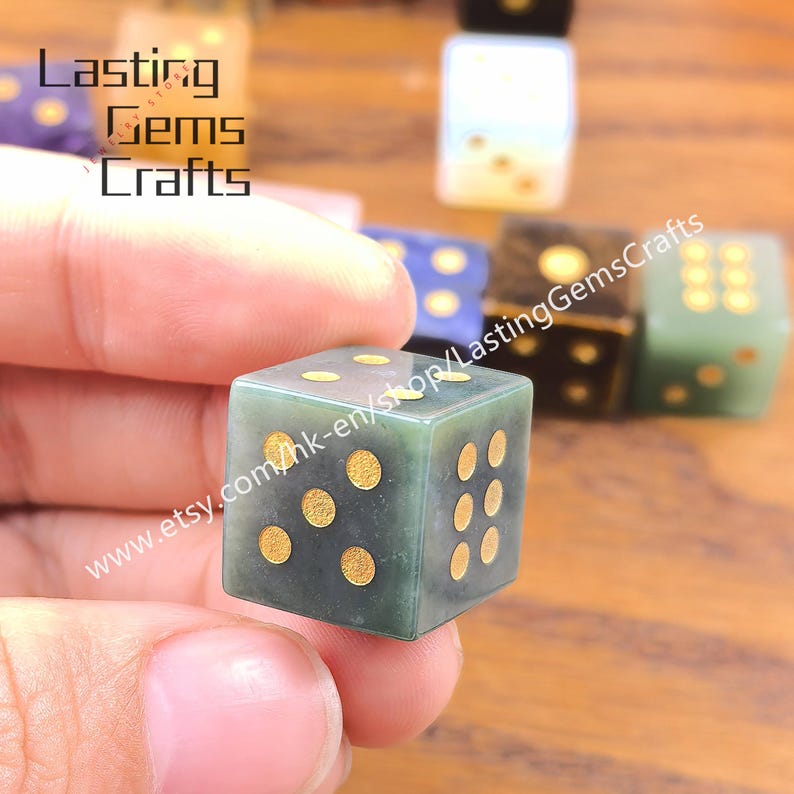 May include: A green gemstone die with gold-colored dots is held in a hand. The die is in focus, with other dice of various colors blurred in the background. The text "Lasting Gems Crafts" and a website address are visible.