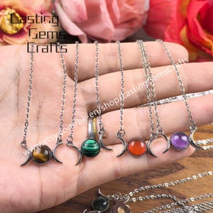 Pode incluir: Uma coleção de colares prateados, cada um com um design de lua crescente e uma pedra preciosa diferente. As pedras preciosas incluem olho de tigre, malaquite, cornalina e ametista. O nome da loja "Lasting Gems Crafts" é visível.