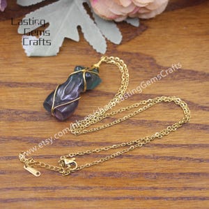 May include: A gold chain necklace with a purple and green fluorite pendant wrapped in gold wire. The pendant is shaped like a human figure. The necklace is on a wooden surface.