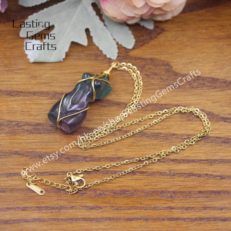 May include: A gold chain necklace with a purple and green fluorite pendant wrapped in gold wire. The pendant is shaped like a human figure. The necklace is on a wooden surface.