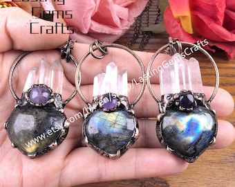 Puede incluir: Tres colgantes en forma de corazón con cristales de cuarzo transparente, detalles de amatista y piedras de labradorita iridiscentes. Los colgantes están suspendidos de aros color cobre. El texto "Lasting Gems Crafts" es visible.