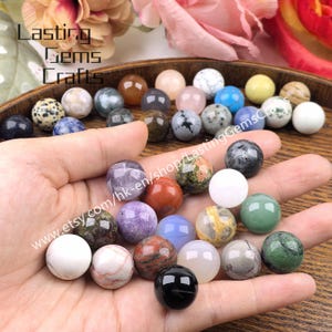 May include: A selection of gemstone spheres in diverse colours and patterns, including white, green, purple, and black. The spheres are displayed in a wooden bowl and in a hand. The text "Lasting Gems Crafts" is visible.