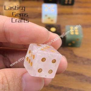 May include: A clear, square dice with gold-colored dots, held in a hand. The dice is translucent, with visible inclusions. Other dice in various colors are visible in the background. The text "Lasting Gems Crafts" and a website address are also visible.