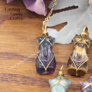 May include: Three gold-wrapped wire-wrapped crystal pendants in the shape of a human figure. The crystals are a dark green, a purple and a brown tiger's eye. The pendants are on a wooden surface. www.etsy.com/hk-en/shop/LastingGemsCrafts