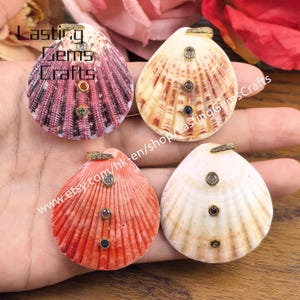 May include: Four seashell pendants featuring gold-toned bails with embedded crystals. Each shell displays a unique color, including deep pink, tan, orange, and cream, with three small, colorful circular gemstones set vertically on the surface.