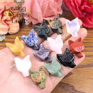 Può includere: Una collezione di figurine di lupi in pietra preziosa scolpite in vari colori, tra cui giallo, blu, rosa e nero. Le figurine sono tenute in una mano, con il testo "Lasting Gems Crafts" visibile sullo sfondo.