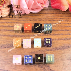 May include: A collection of gemstone dice in various colors, including pink, blue, black, and green, with gold-colored dots. The dice are arranged on a wooden surface, with text that reads "Lasting Gems Crafts" and a website address.