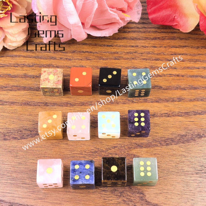 May include: A collection of gemstone dice in various colors, including pink, blue, black, and green, with gold-colored dots. The dice are arranged on a wooden surface, with text that reads "Lasting Gems Crafts" and a website address.