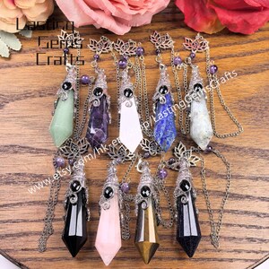 May include: A collection of gemstone pendant necklaces in various colors, including green, purple, white, blue, black, pink, and gold. Each pendant is suspended from a silver-colored chain and features a decorative top with a black stone accent. The pendants are displayed on a wooden surface.