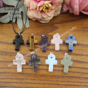 May include: An assortment of eight Ankh pendants made from various gemstones, including black onyx, tiger's eye, amethyst, rose quartz, clear quartz, blue lace agate, and green aventurine. The pendants are displayed on a wooden surface.