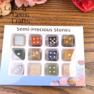 May include: A set of twelve semi-precious stone dice in a white box. Each die has a different color and pattern, with gold-colored dots indicating the numbers. The box is labeled "Semi-Precious Stones."