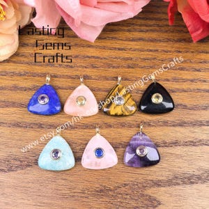 May include: A collection of triangular gemstone pendants in various colors, including blue, pink, and black, each with a small, central gemstone. The pendants are displayed on a wooden surface. The text "Lasting Gems Crafts" is visible.