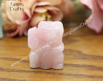 May include: A small, pink, carved rose quartz figurine of a beckoning cat, also known as a Maneki-Neko. The figurine is sitting on a wooden surface.