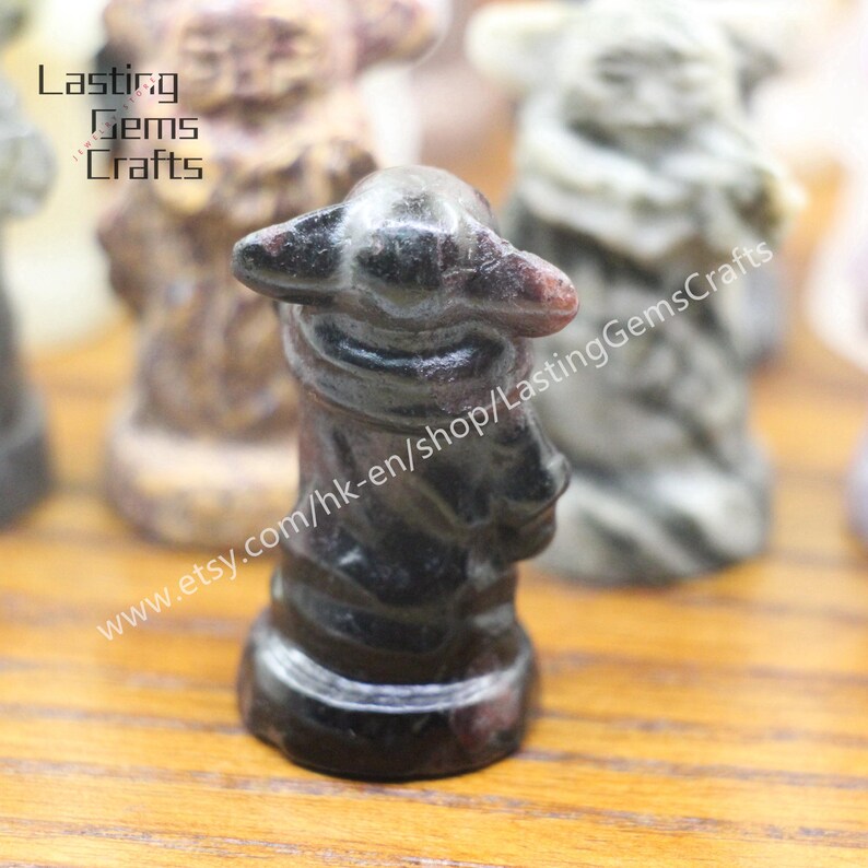 May include: A small, dark gray and black stone figurine of a person wearing a hat. The figurine is carved from a single piece of stone and has a detailed face and clothing.